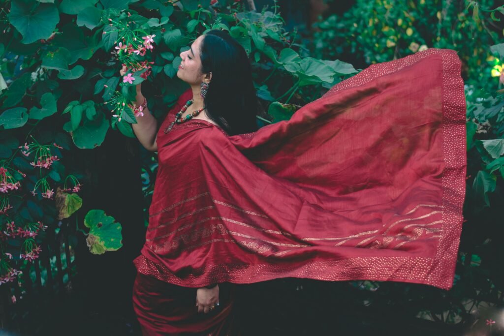 Woman in a flowing red saree stands among lush green foliage, embracing traditional Indian beauty.
